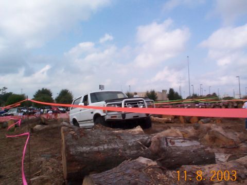 1992 Ford Bronco - A white SUV navigates a challenging obstacle course made of logs and dirt, with other vehicles visible in the distance.