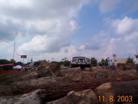 1992 Ford Bronco - SUVs tackle a challenging off-road course marked by logs and rocks under a cloudy sky.