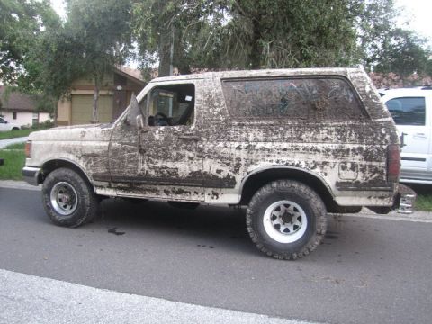 1988 Ford Bronco - A heavily mud-caked 1987 Ford Bronco is parked on a residential street next to another vehicle.