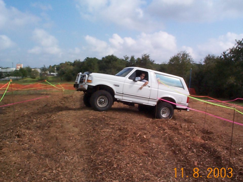 White Ford Bronco with brush guard climbing a dirt mound in a ribbon-marked off-road course