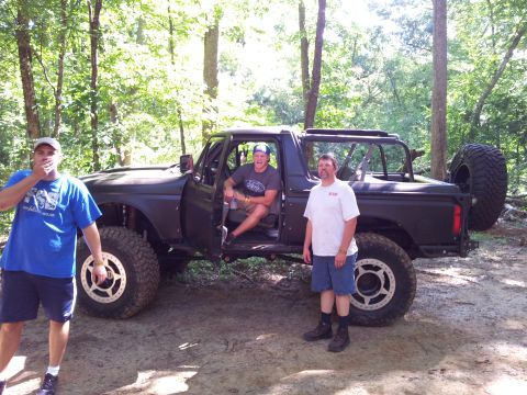 2012 Ford Bronco - A dark-colored SUV with lifted suspension and large tires is parked in a wooded area with three men standing around it.