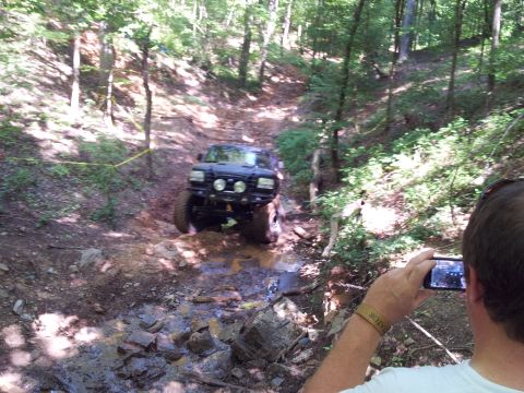 2012 Ford Bronco - A person films an off-road vehicle as it maneuvers through a challenging, water-filled dirt path in a forest.