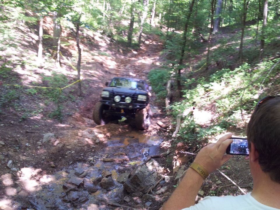 Black off-road SUV driving through a muddy, wooded trail with large tires and extra lights on the front