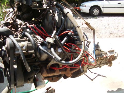 1995 Ford Bronco - A vehicle engine and transmission assembly is shown, suspended by chains, with a white car visible in the background.