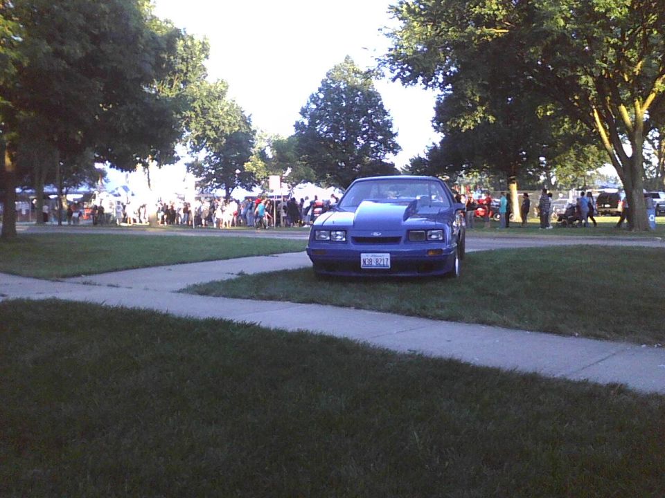 Blue Ford Mustang with a large hood scoop parked on grass near a crowd of people