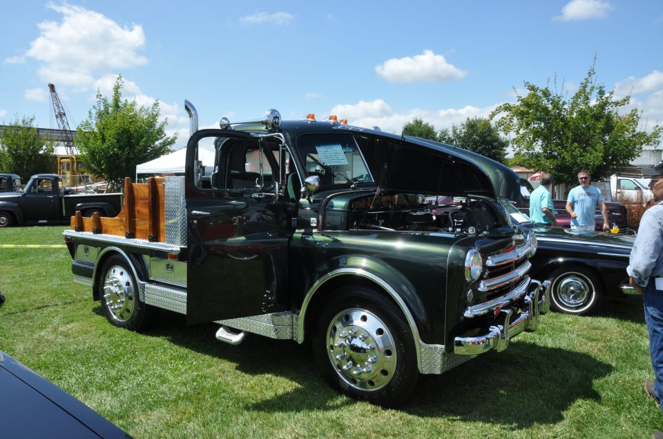 Green flatbed truck with wooden stake sides, chrome grille, and hood open. Another car is parked next to it