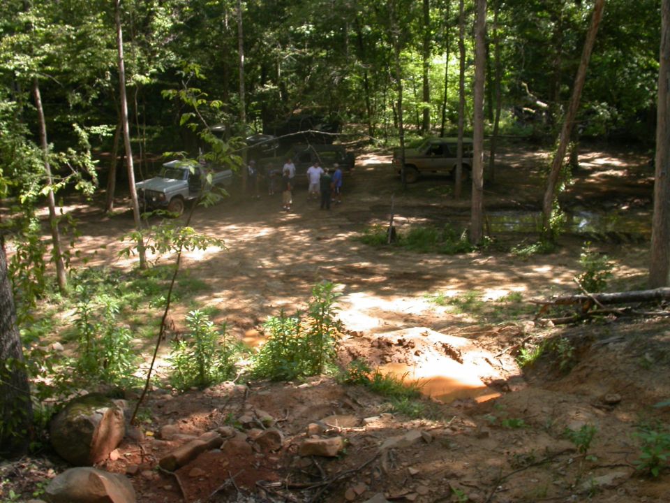 Two off-road vehicles are parked in a dirt clearing surrounded by trees, with several people standing nearby