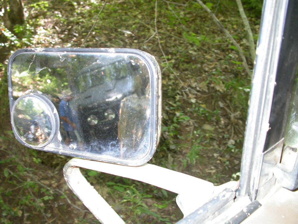 Reflection of a dark SUV in a dirty side mirror, showing round headlights