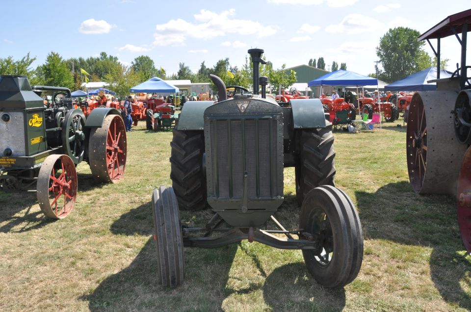 Front view of a dark green tractor with large rear wheels and smaller front wheels, parked on grass with other tractors and tents in the background
