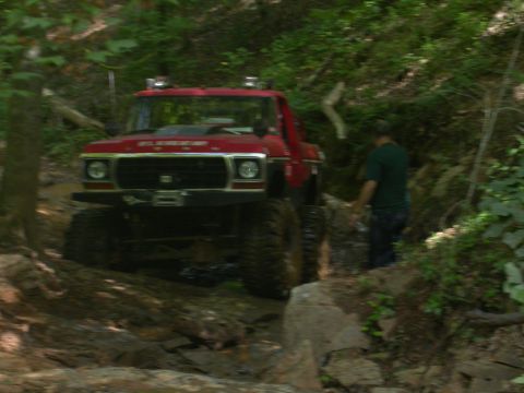 1990 Ford Bronco - A red off-road truck navigates a challenging, wooded trail with large rocks and mud.