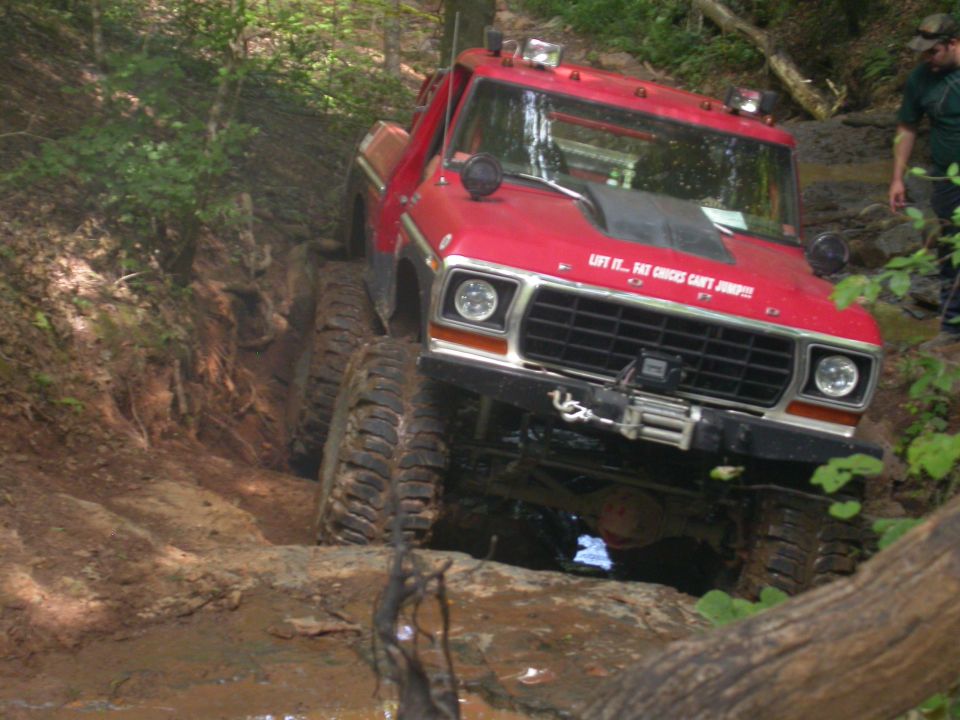 Red off-road truck with large tires navigating a muddy trail. Text on hood says 'LIFT IT... FAT CHICKS CAN'T JUMP!!!'