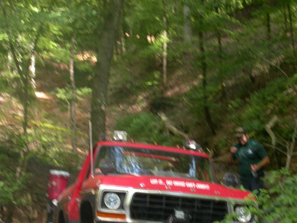 Red truck with a white stripe on the hood, off-road lights, and a person standing next to it in a wooded area