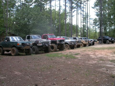 1990 Ford Bronco - A row of modified Ford Broncos and trucks are parked in a dirt lot surrounded by trees.