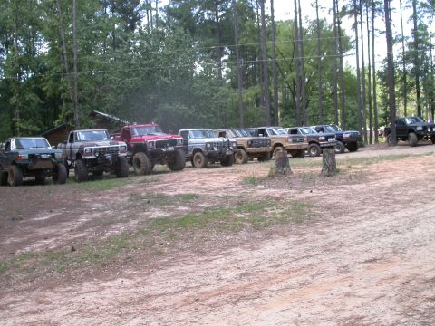 1990 Ford Bronco - A group of modified Ford Broncos and trucks are parked in a dirt lot surrounded by trees.