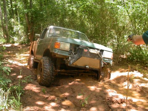 1990 Ford Bronco - A modified truck navigates a muddy trail through a wooded area.