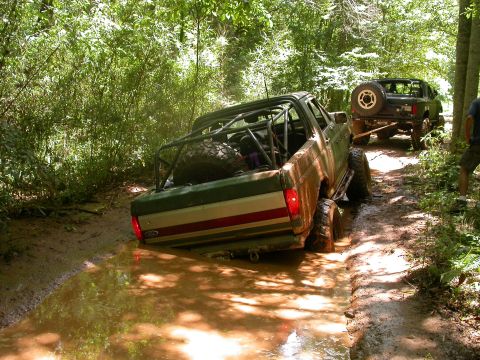 1990 Ford Bronco - Two off-road vehicles navigate a challenging muddy path through a dense forest.