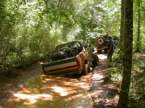 1990 Ford Bronco - An off-road pickup truck with a roll cage is stuck in a muddy trail, being pulled by another vehicle.