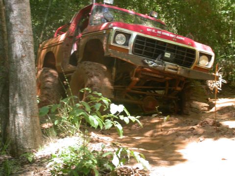 1990 Ford Bronco - This heavily modified SUV is deep in the mud on a wooded trail, with its front end lifted.