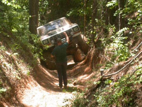 1990 Ford Bronco - A man observes a white Ford Bronco as it maneuvers through a challenging off-road trail in a forest.