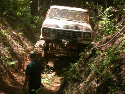 1990 Ford Bronco - A man stands by a modified Ford Bronco as it climbs out of a muddy, wooded trail.