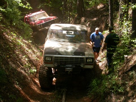 1990 Ford Bronco - Two modified 4x4s navigate a challenging off-road trail through a wooded area.