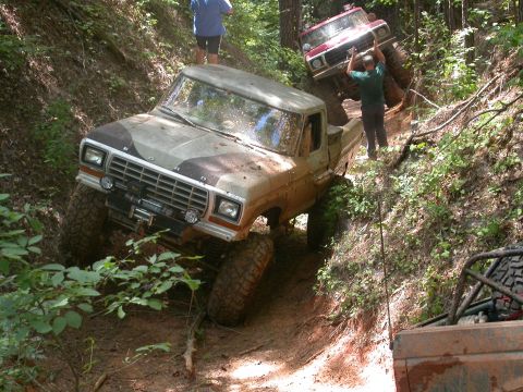 1990 Ford Bronco - Two off-road trucks navigate a challenging dirt trail, one with a winch and camouflage paint, the other red and climbing the incline.