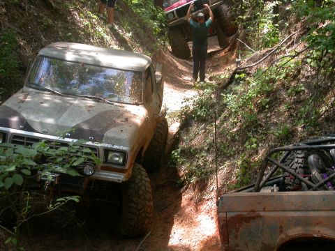 1990 Ford Bronco - Several off-road vehicles navigate a challenging, muddy trail in a wooded area.