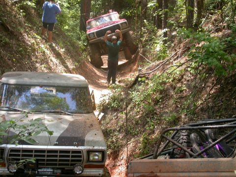 1990 Ford Bronco - Multiple off-road vehicles navigate a steep, wooded trail with assistance from onlookers.