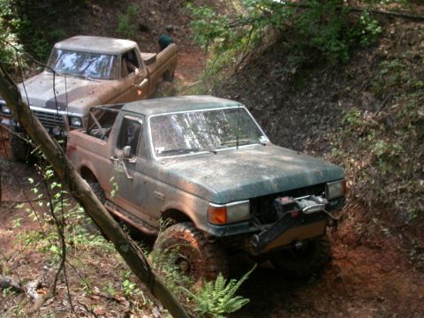 1990 Ford Bronco - Two pickup trucks navigate a muddy, wooded trail.