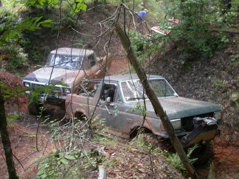 1990 Ford Bronco - Two 4x4 vehicles navigate a challenging, muddy off-road course surrounded by dense foliage.