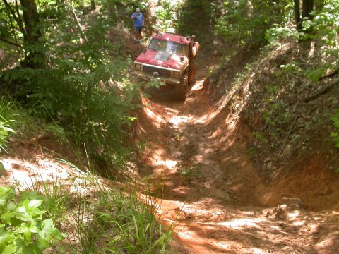1990 Ford Bronco - A red off-road vehicle navigates a steep, muddy trail surrounded by trees.