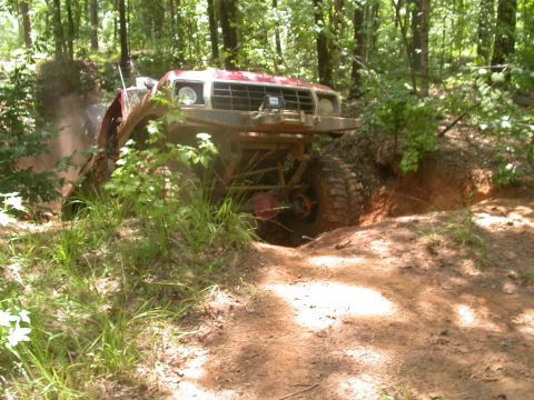 1990 Ford Bronco - A red off-road vehicle navigates a challenging dirt trail through a forest.