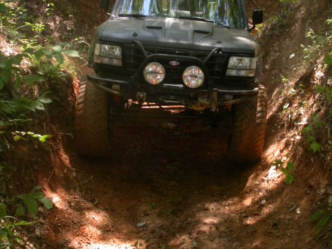 1990 Ford Bronco - An off-road SUV navigates a tight, muddy trail surrounded by dense forest foliage.