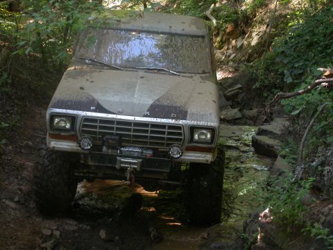 1990 Ford Bronco - This heavily muddied 1980 Ford Bronco navigates a challenging off-road course with rocky terrain and water obstacles.