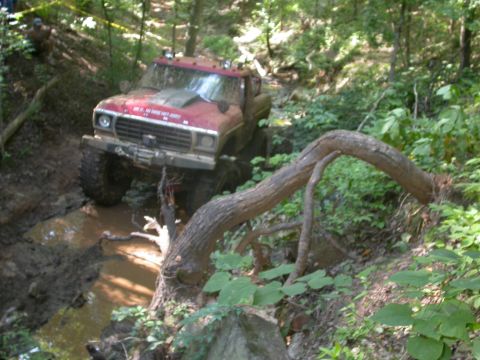 1990 Ford Bronco - This modified 1979 Ford F-Series truck tackles a challenging off-road course, navigating through a muddy stream and dense foliage.