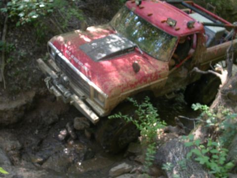 1990 Ford Bronco - This red truck is navigating a difficult off-road trail, stuck in mud and rocks.