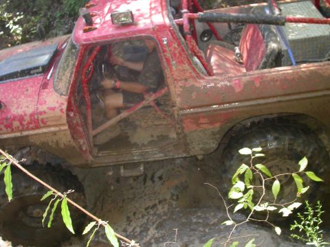 1990 Ford Bronco - A heavily modified red off-road vehicle navigates a muddy trail, its interior protected by a roll cage.