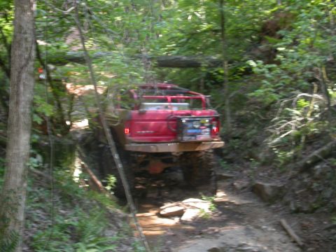 1990 Ford Bronco - A red SUV is shown traversing a challenging off-road trail through a dense forest.