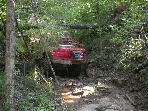 1990 Ford Bronco - A red off-road SUV is shown traversing a challenging, rocky path through a dense forest.