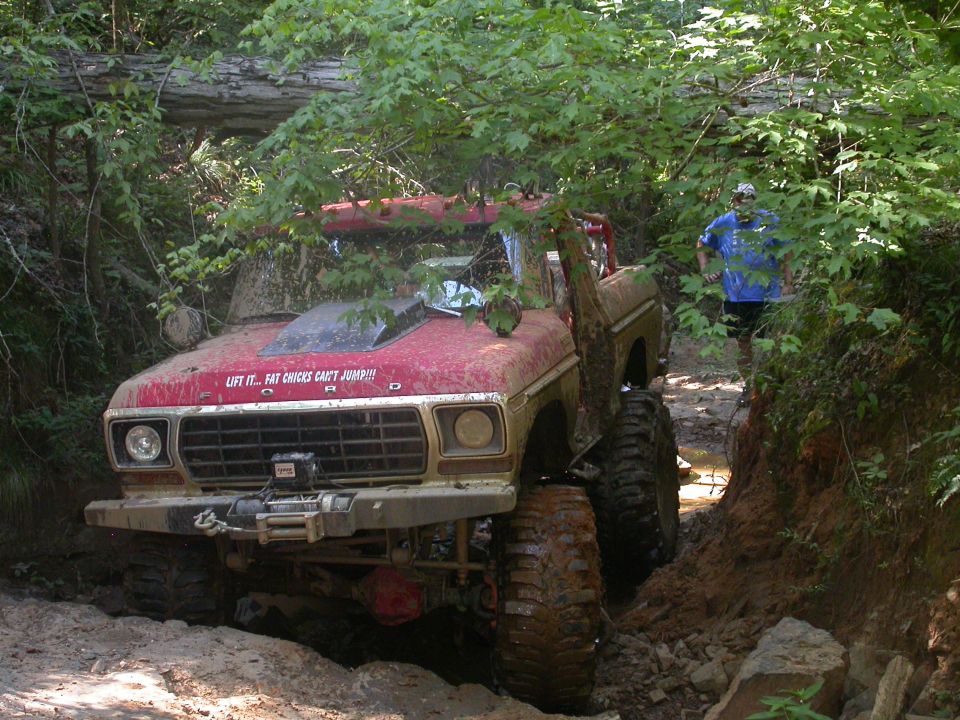 Modified red Ford Bronco with large tires stuck in a muddy trail, text on hood reads 'LIFT IT... FAT CHICKS CAN'T JUMP!!!'