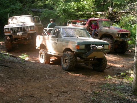 1990 Ford Bronco - Several off-road vehicles are parked on a dirt path in a wooded area.
