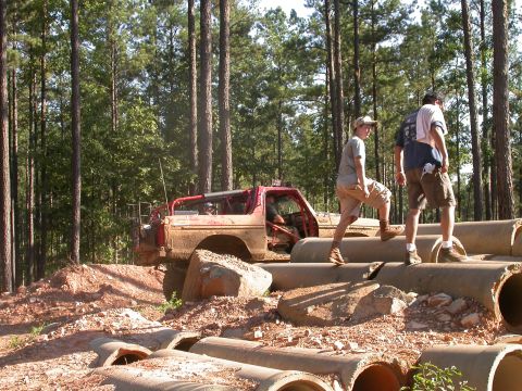 1990 Ford Bronco - A modified red off-road vehicle navigates a challenging obstacle course in a wooded area.