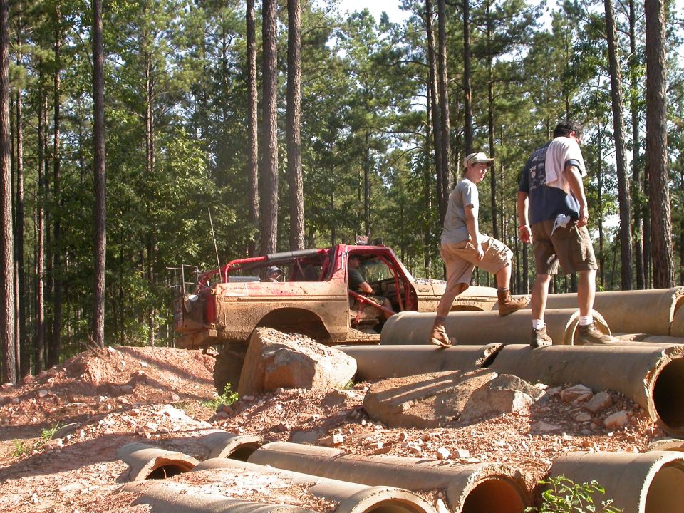 Mud-covered red off-road vehicle with roll cage, driving over concrete pipes