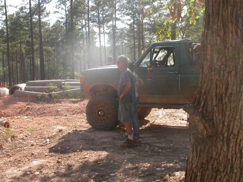 1990 Ford Bronco - A dark green 1990 Ford Bronco with lifted suspension and large tires is parked on a dirt road in a wooded area.