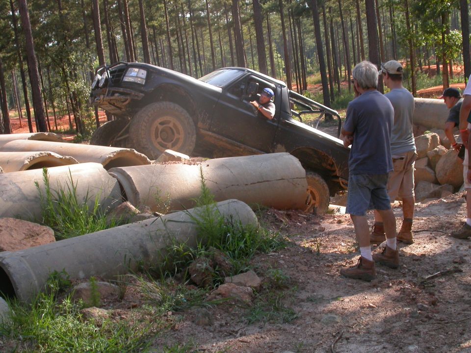 Black off-road truck driving over large concrete pipes in a wooded area with people watching