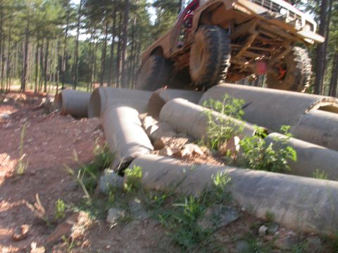 1990 Ford Bronco - A lifted off-road vehicle navigates a challenging obstacle course of large concrete pipes in a forest setting.