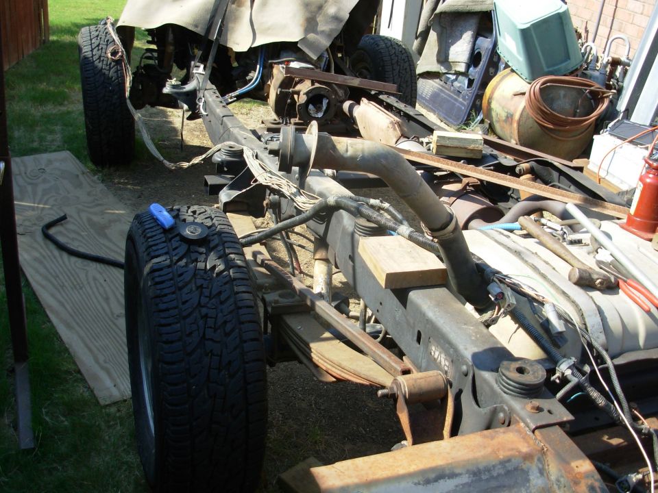 Bare truck frame with leaf springs, exhaust and loose wiring harness, tire leaning against it outdoors