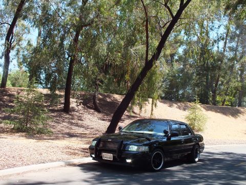 2003 Ford Crown Victoria - Distant front view of a black Mercury sedan on a road bordered by trees and a dry hillside.