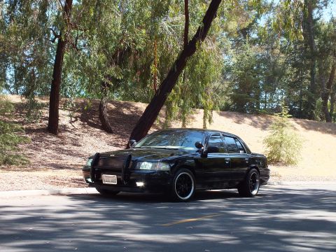 2003 Ford Crown Victoria - Front-left view of a lowered black sedan on staggered dark wheels in shade.