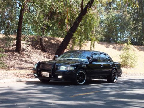 2003 Ford Crown Victoria - Front-quarter view of a lowered black sedan with dark staggered wheels in shade.